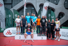 A group of participants of a sports event is standing on and in front of the podium in a sports hall during a triathlon ceremony. Behind them, flags and banners of the Polish Triathlon Association and AZS are visible.