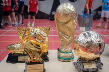 Zestaw sportowych trofeów ustawionych na stole: złota statuetka bramkarza, puchar w kształcie Złotej Piłki dla MVP oraz duży puchar przypominający trofeum mistrzostw świata; w tle widać uczestników turnieju na hali sportowej.