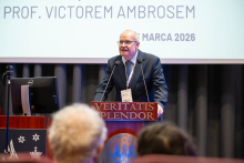 A person standing at a podium and speaking during an event, with a presentation slide in the background showing the date ‘13 March 2026’ and part of the session title.
