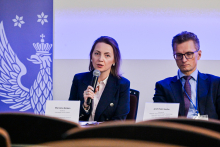 Two people are sitting at a table during a conference or panel discussion. The person on the left is holding a microphone. In front of her is a nameplate reading “Marzena Nielsen.” On the right sits a man with a nameplate reading “Prof. Piotr Socha.” On the left side of the image, a blue banner with a white eagle and stars is visible.