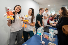 Female participants standing at a booth, presenting plush mascots, next to medical materials, leaflets, and other informational items; in the background, people can be seen conversing at neighboring stations.