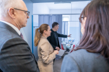 Several people are standing in a hospital room, listening to a man who is pointing toward the equipment located behind a curtain; a medical bed and a bright window are visible in the background.