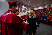 A woman in an academic gown approaches a group of three professors and holds a document. She extends her hand toward the professor standing closest to her. The professor reciprocates the gesture.