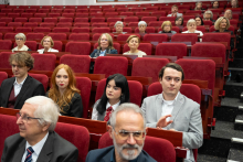 Participants of the event are seated in the red armchairs of the large auditorium, observing the course of the ceremony; some seats remain unoccupied.