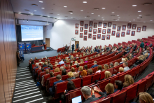 The participants are seated in multiple rows of red chairs and are listening to a speech given at a podium on the left side of the room. The presenter is standing next to a screen displaying a conference slide. In the lower part of the room, people can be seen taking notes or holding devices, and numerous portraits hang on the wall to the right.