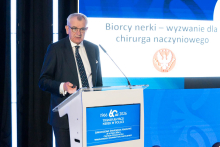 A speaker stands at the lectern, delivering a presentation. He looks toward the large screen, which displays a title regarding challenges in vascular surgery in kidney recipients. One hand rests on the podium, while the other holds a clicker or notes. Next to the speaker is a blue screen displaying information about the jubilee symposium.
