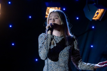 A woman is standing on stage and singing into a microphone. She is holding the microphone with one hand and making a relaxed sideways gesture with the other. Her posture is confident, and her vocal engagement is visible in her expression and body position. Behind her, small blue lights are shining.