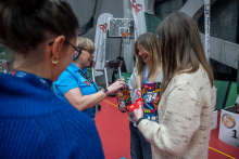 Several people are standing together in a sports hall, holding colourful collection cans related to the 34th Grand Finale of the Great Orchestra of Christmas Charity. One person in a blue T‑shirt is pointing at something on a can, while the others listen.