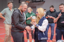 Podczas ceremonii w hali sportowej kobieta wręcza zawodnikowi nagrodę w formie złotej piłki, jednocześnie ściskając jego dłoń; wokół stoją inni uczestnicy wydarzenia.