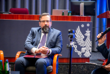 A person in a navy-blue suit is sitting in an armchair during a panel discussion, holding documents on their lap. Next to them, the microphone of another participant is visible, and in the background there is a lectern with an emblem depicting an eagle.