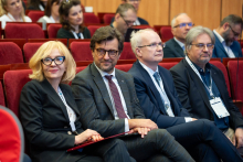 A group of people seated in an auditorium on red chairs during a formal event or conference, dressed in business attire and listening to a presentation