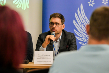 A middle‑aged man, dressed formally, is sitting at a table and speaking into a microphone. In front of him is a nameplate reading “dr n. med. Maciej Janik” along with information about his affiliation with the European Reference Network Adult LCH, UZL. In the background, blue‑and‑white patterns featuring wing and star motifs are visible. In the foreground, blurred silhouettes of other people can be seen.
