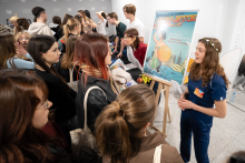 A person presenting a poster with a colorful illustration during an educational event, talking with a group of participants gathered around the booth.