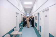 A group of people is standing in a long, bright hospital corridor, talking during a visit; one person is pointing further down the corridor while the others listen.
