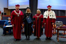 Four people stand together during the ceremony. The graduate in an academic gown stands in the middle, holding a document in front of her. On both sides of her stand professors in ceremonial gowns, looking toward the photographer.