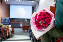 A bouquet of pink roses rests on the lap of a person sitting in one of the front rows. In the background, the speaker is standing at the podium and giving a presentation. Participants are seated in rows of red chairs and listening to the presentation displayed on a large screen.