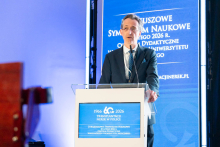 A speaker stands at the lectern, delivering a presentation during the symposium. He rests his hands on the podium and looks toward the audience. Next to him is a tall blue screen displaying information about the jubilee event.