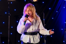 A woman is standing on stage and singing into a microphone. She is holding it close to her mouth and making a decisive gesture to the side with her other hand. Her posture is confident, and her facial expression suggests engagement in the performance.