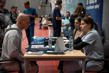 A man is sitting at a small table and talking to a woman dressed in medical attire. On the tabletop there are informational materials and oral hygiene products. In the background, other people participating in the event in the sports hall can be seen.