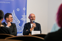 Two people are sitting at a table during a panel discussion. The person on the right is holding a microphone and speaking, while the person on the left is listening. Behind them, a blue banner with the white inscription “WARSZAWSKI UNIWERSYTET MEDYCZNY” and an eagle with outstretched wings is visible. In front of the speaker lies a nameplate reading “Mariusz Jop.”