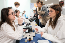People working at a laboratory station, examining a specimen under a microscope and discussing the results, with reagents and auxiliary materials visible on the table.