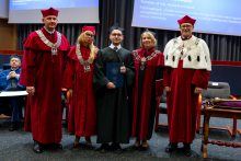Five people stand together during the ceremony. The graduate in an academic gown holds a document and stands in the middle of the group. On both sides of him stand professors in ceremonial gowns. All of them are looking toward the photographer.