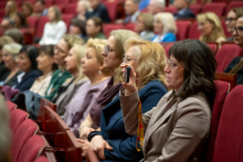 A few people are sitting in red chairs and listening to a speech. In the foreground, a woman raises her phone and points it toward the stage. Next to her, other attendees are seated with their hands resting on their laps or clasped together. In the further rows, a larger group of participants can be seen taking their seats in the auditorium.