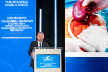 A speaker stands at the lectern, delivering a presentation. One hand rests on the podium, while the other is positioned near the microphone. He looks toward the audience. Next to the speaker stands a tall blue screen displaying information about the jubilee symposium. On the right side, a large slide is visible, showing a stage of an operation with the organ and the hands of the person performing the procedure.