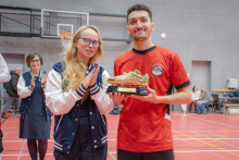 A player in a red jersey poses with a Golden Boot award, holding the trophy in his hands, while a person standing next to him claps appreciatively in the sports hall.