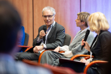 People seated on stage during a discussion panel, holding microphones and participating in a conversation in a professional setting.