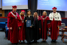 Six people stand together during the ceremony. The graduate and her supervisor, both in academic gowns, stand in the center holding a document. Professors in ceremonial gowns stand on either side of them. All of them are looking toward the photographer.