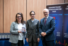 Three people are standing next to each other against the backdrop of roll‑ups from the University Clinical Center of the Medical University of Warsaw. The woman on the left is holding an open box with a medal. Next to her stand a woman and a man dressed in formal attire. All three are looking toward the camera.