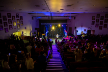 The audience is standing and sitting in a large hall, watching a performance on a brightly lit stage. One person is standing in the center of the stage, singing or speaking into a microphone. The audience is looking toward the stage, and some are recording the event with their phones.