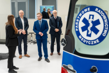 A group of people in elegant attire is standing in a training room, listening to a presentation led by an instructor. In the foreground, a part of an ambulance with the ‘Państwowe Ratownictwo Medyczne’ logo is visible.