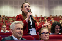A person standing between rows of red auditorium seats asks a question during a conference, holding a microphone, surrounded by a large audience