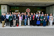 Around 30 people are standing in front of a building entrance. They are dressed formally or semi‑formally, and some are wearing lanyard ID badges. In the background, a wall with a large emblem and two signs on either side is visible.