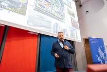 A person delivering a presentation in a lecture hall, standing by a microphone in front of a large screen displaying architectural visualizations of a building complex.