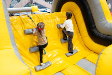 Two children are climbing soft steps on a yellow‑and‑gray inflatable climbing wall in a sports hall.
