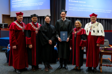 Six people stand together during the ceremony. The graduate in an academic gown stands in the middle holding a document. On both sides of him stand two people in graduation gowns and three professors in ceremonial gowns. All of them are looking toward the photographer.