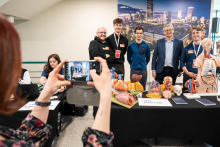 A person taking a photo of a group presenting anatomical models arranged on a table during an educational event; behind the table, participants are displaying various organ models against the backdrop of a large city panorama photograph.
