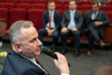 A person holding a microphone is speaking during the discussion, seated in the front row of the auditorium, facing a group of event participants