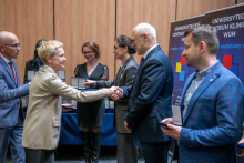 Six people are standing in two rows and taking part in an award ceremony. A man in a suit is shaking hands with the person standing opposite him. Several participants are holding cases with medals. In the background, a table with neatly arranged cases and roll-ups of the University Clinical Center of the Medical University of Warsaw can be seen.