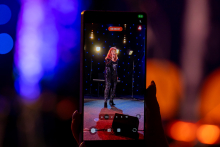 A phone held in a hand is recording a performance on stage. On the screen, a woman can be seen singing into a microphone, standing in the spotlight. Her posture is confident, and her gestures highlight her engagement in the performance. In the background, small blue lights are shimmering.