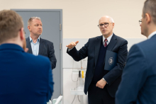 A person in an elegant suit is gesturing while speaking in a training room, surrounded by several other people standing in a semicircle.
