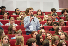 A person standing between rows of red auditorium seats asks a question during a conference, holding a microphone, surrounded by a large audience