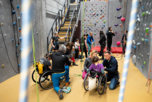 A group of people is participating in climbing activities on a climbing wall in a sports hall. Several people in wheelchairs are preparing for belaying or climbing with the assistance of instructors. Others are standing by the ropes or under the wall, checking the equipment. In the background, colourful climbing holds and stairs leading to an upper level of the facility are visible.