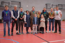 Grupa uczestników stoi razem na boisku w hali sportowej, część z nich ma na szyjach medale, a w pierwszym rzędzie stoją również dzieci biorące udział w ceremonii.