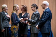 Five people are standing in a small group during an award ceremony. The two women in the center are holding medal cases and handing them to each other, making hand gestures. Next to them, other individuals are standing and observing the exchange. In the background, wooden panels and a row of closed cases arranged on a table can be seen.