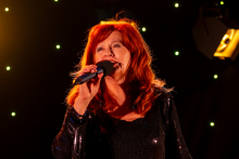 A woman is standing on stage and singing into a microphone. She is holding it close to her mouth, and with her other hand she is making a decisive sideways gesture. Her posture is confident, and her facial expression suggests engagement in the performance. In the background, small green lights are shining.