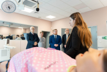 A group of people in elegant attire is standing in a medical simulation room, listening to a presentation by an instructor. In the foreground, a training mannequin wearing a pink T‑shirt is visible.